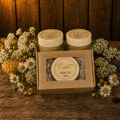 Two jars of Under Eye salve with a box in front on a wooden surface with flowers around.